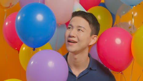 Studioporträt-Eines-Mannes-Mit-Partyhut,-Der-Seinen-Geburtstag-Feiert-Und-Sich-Hinter-Luftballons-Versteckt